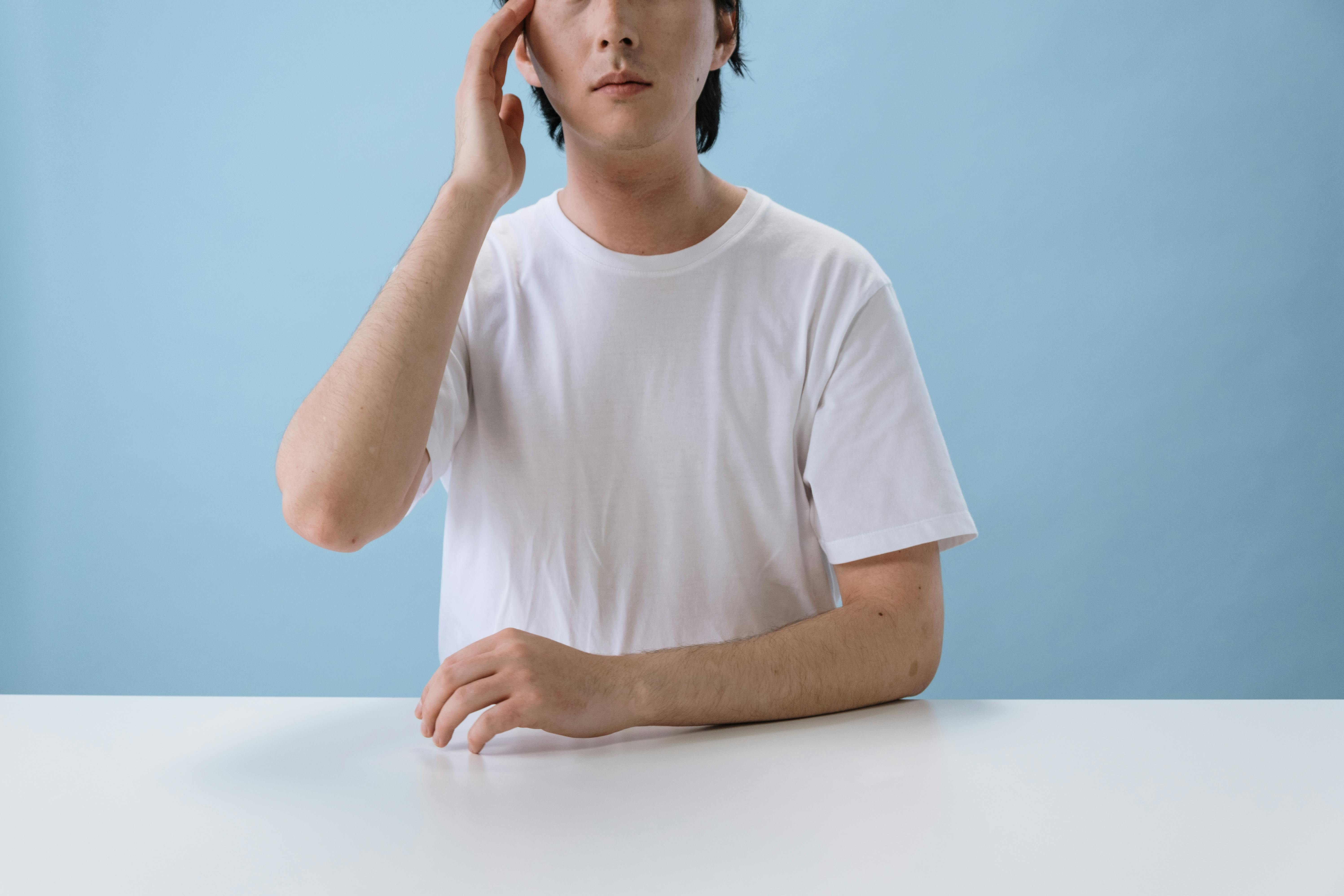 A man in a blank white t-shirt sits pensively against a blue background.